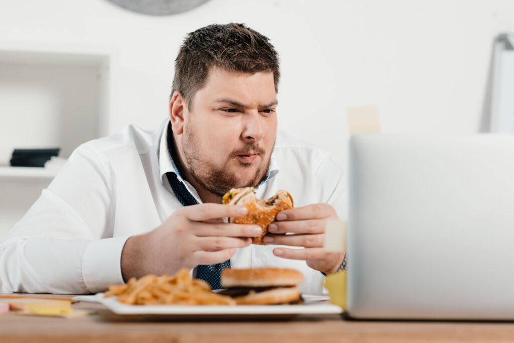 Overweight businessman working while eating hamburger and french fries in office